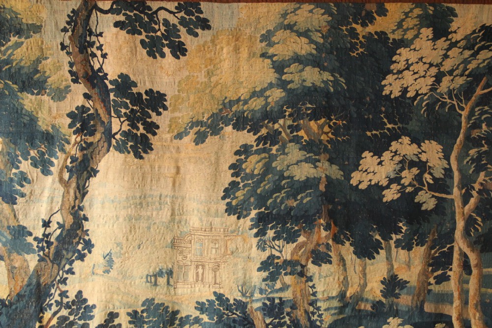 A large woven Flemish verdure tapestry featuring a dense forest landscape with trees, botanical plants, a central chateau view, and a decorative foliage border. View X