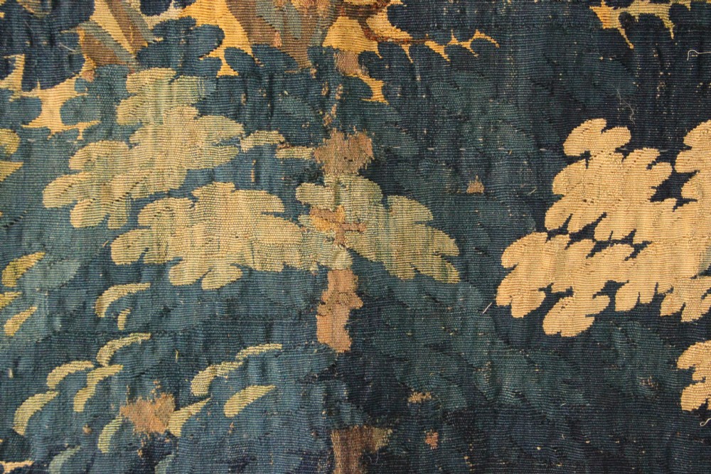 A large woven Flemish verdure tapestry featuring a dense forest landscape with trees, botanical plants, a central chateau view, and a decorative foliage border. View XI