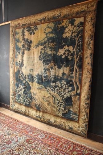 A large woven Flemish verdure tapestry featuring a dense forest landscape with trees, botanical plants, a central chateau view, and a decorative foliage border. View II