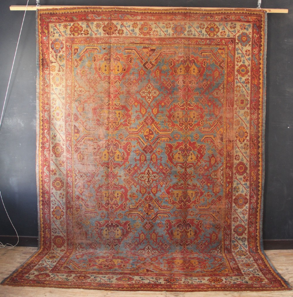 A large antique Turkish Oushak wool carpet featuring a pale blue field layered with terracotta and gold medallion motifs and a patterned border.