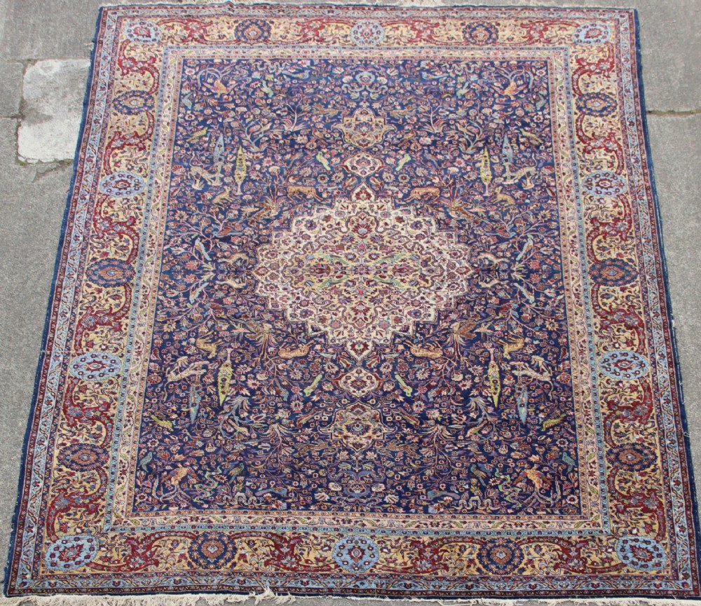 A large hand-knotted Persian Tabriz carpet featuring a central cream medallion surrounded by a navy blue field with intricate floral and hunting motifs, framed by a detailed red border.