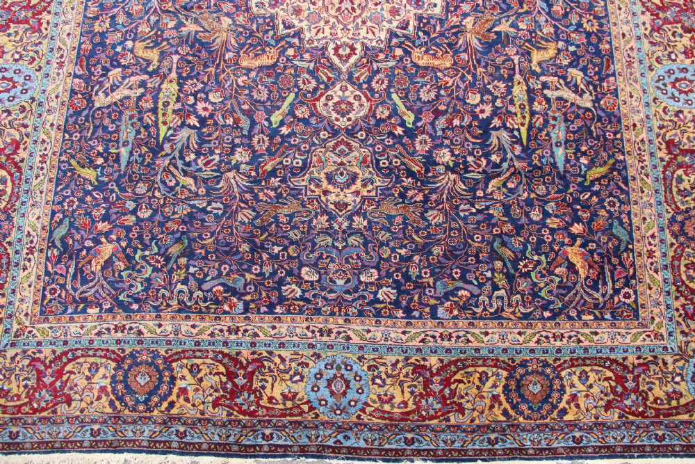 A large hand-knotted Persian Tabriz carpet featuring a central cream medallion surrounded by a navy blue field with intricate floral and hunting motifs, framed by a detailed red border. View II
