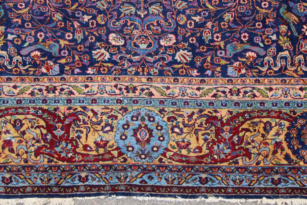 A large hand-knotted Persian Tabriz carpet featuring a central cream medallion surrounded by a navy blue field with intricate floral and hunting motifs, framed by a detailed red border. View III