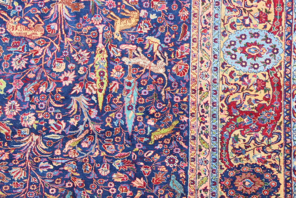 A large hand-knotted Persian Tabriz carpet featuring a central cream medallion surrounded by a navy blue field with intricate floral and hunting motifs, framed by a detailed red border. View IV