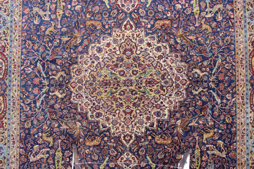 A large hand-knotted Persian Tabriz carpet featuring a central cream medallion surrounded by a navy blue field with intricate floral and hunting motifs, framed by a detailed red border. View V