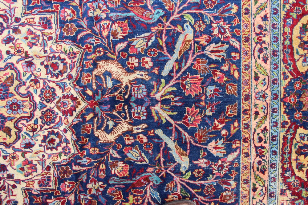 A large hand-knotted Persian Tabriz carpet featuring a central cream medallion surrounded by a navy blue field with intricate floral and hunting motifs, framed by a detailed red border. View VI