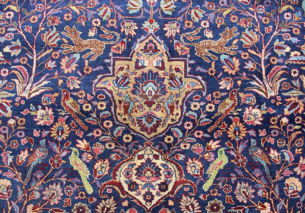 A large hand-knotted Persian Tabriz carpet featuring a central cream medallion surrounded by a navy blue field with intricate floral and hunting motifs, framed by a detailed red border. View IX