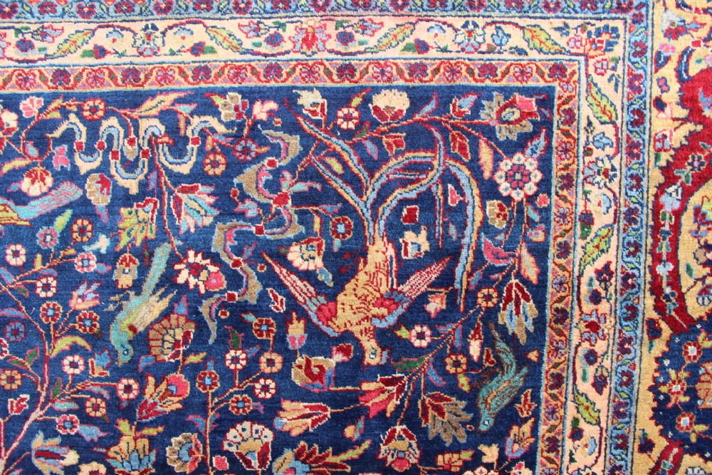 A large hand-knotted Persian Tabriz carpet featuring a central cream medallion surrounded by a navy blue field with intricate floral and hunting motifs, framed by a detailed red border. View X