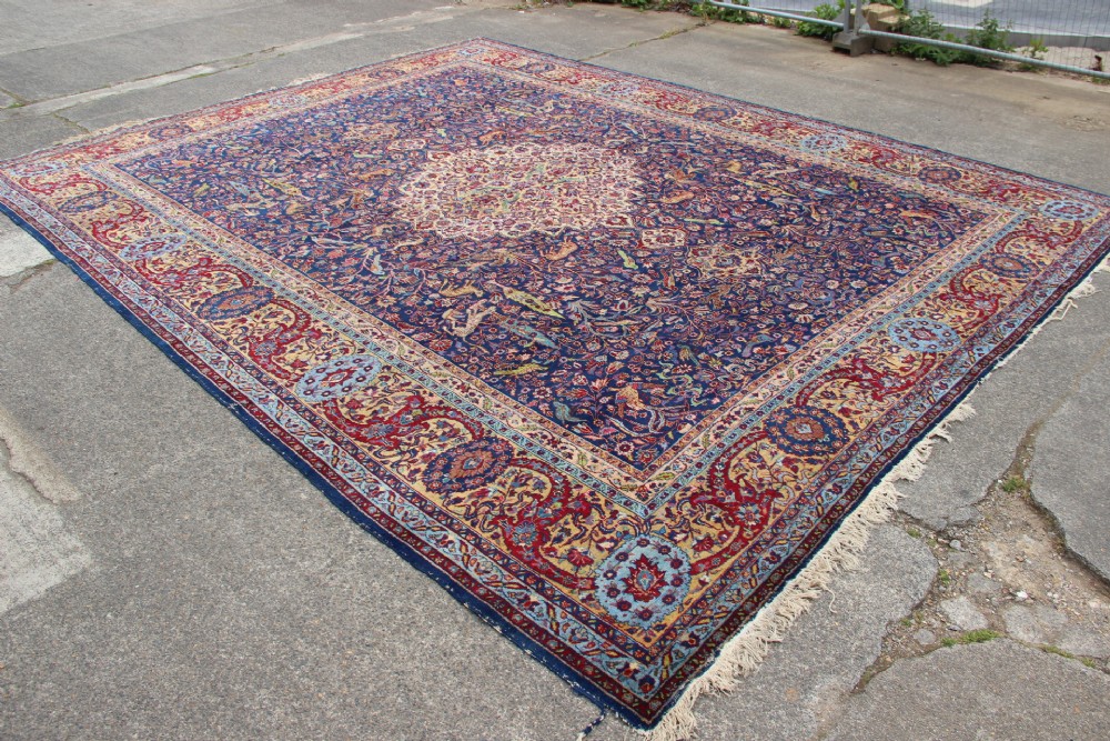 A large hand-knotted Persian Tabriz carpet featuring a central cream medallion surrounded by a navy blue field with intricate floral and hunting motifs, framed by a detailed red border. View XII