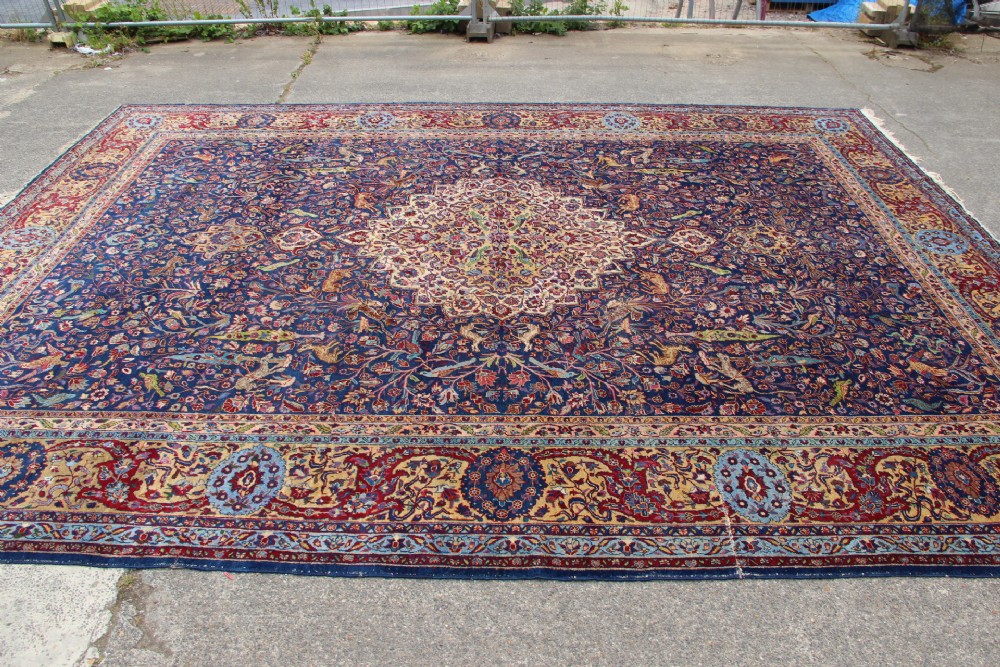 A large hand-knotted Persian Tabriz carpet featuring a central cream medallion surrounded by a navy blue field with intricate floral and hunting motifs, framed by a detailed red border. View XIII