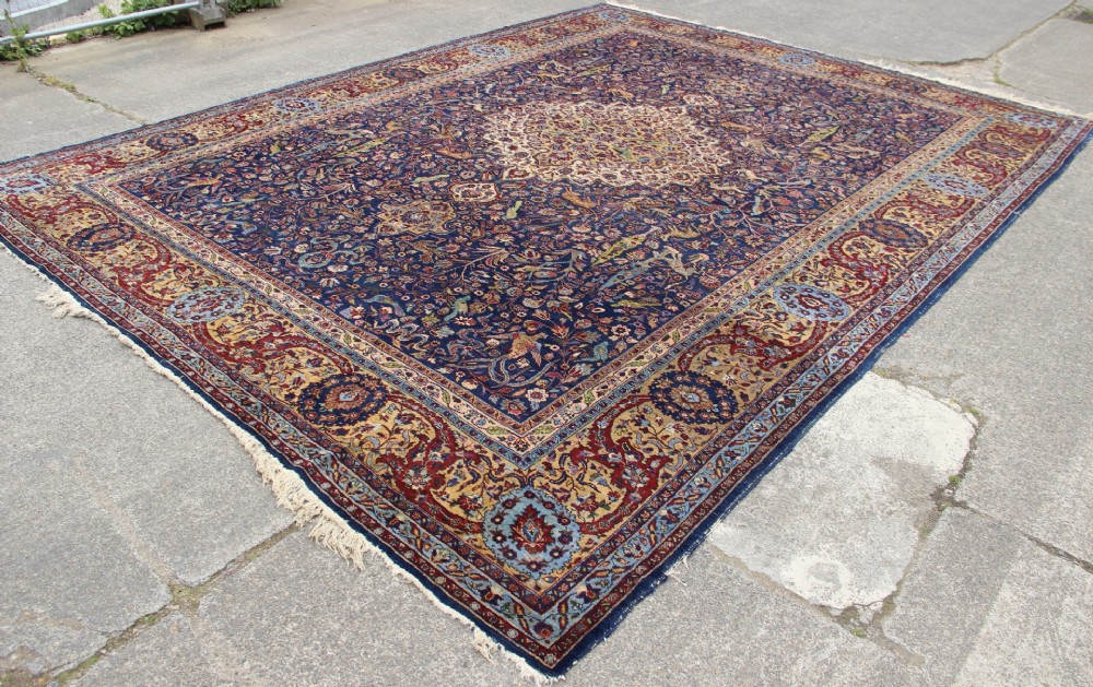 A large hand-knotted Persian Tabriz carpet featuring a central cream medallion surrounded by a navy blue field with intricate floral and hunting motifs, framed by a detailed red border. View XIV