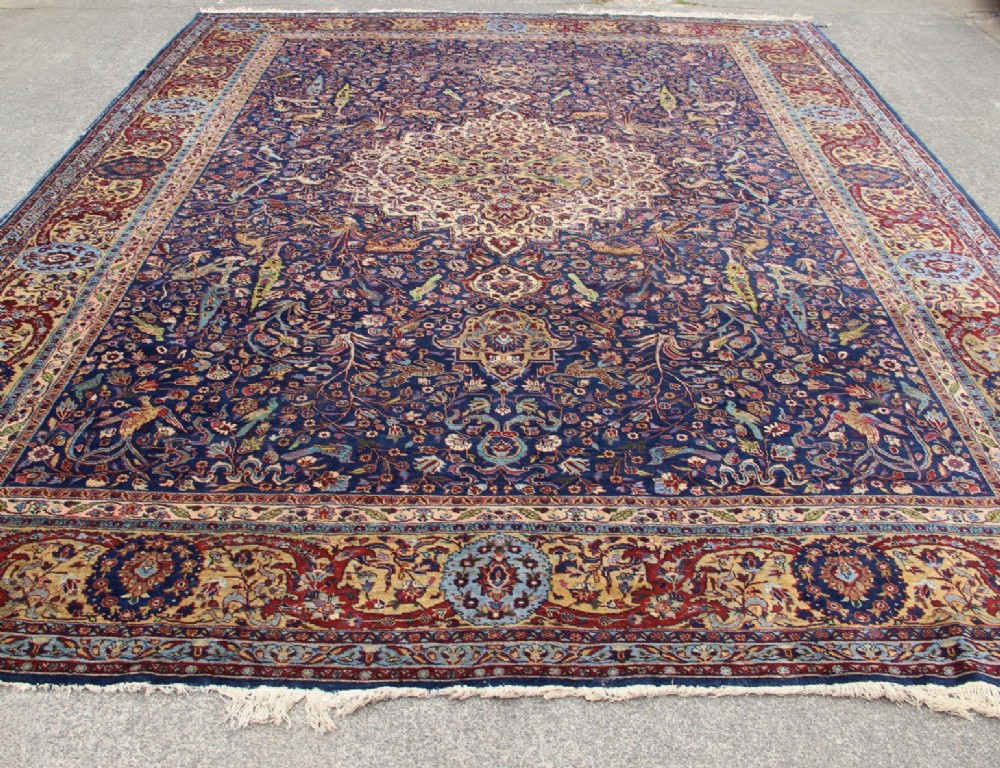 A large hand-knotted Persian Tabriz carpet featuring a central cream medallion surrounded by a navy blue field with intricate floral and hunting motifs, framed by a detailed red border. View XV