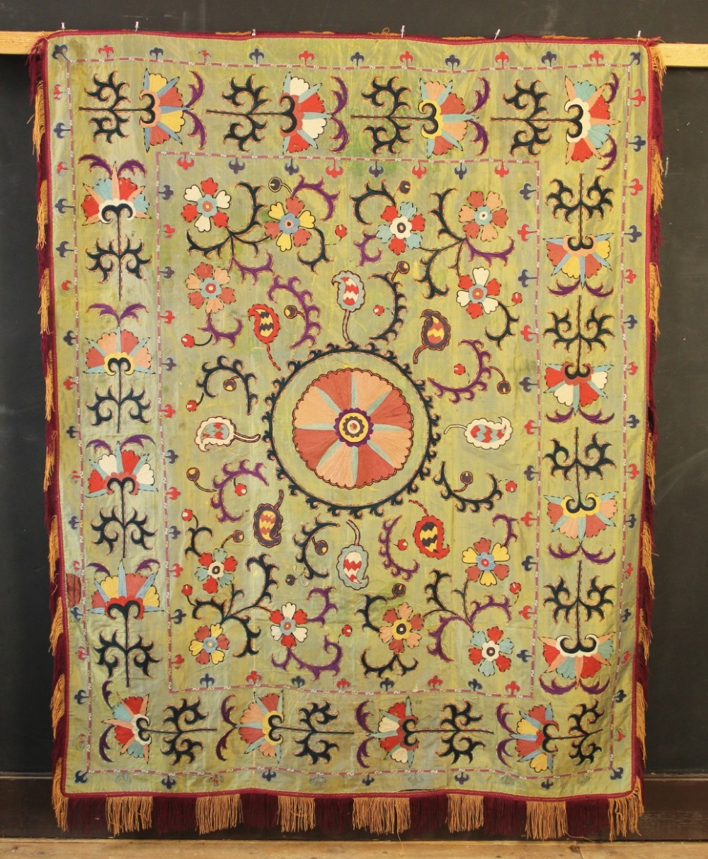 A rectangular green silk embroidered Suzani panel featuring a central circular solar motif flanked by intricate scrolling vines, botanical borders, and fringe edging.