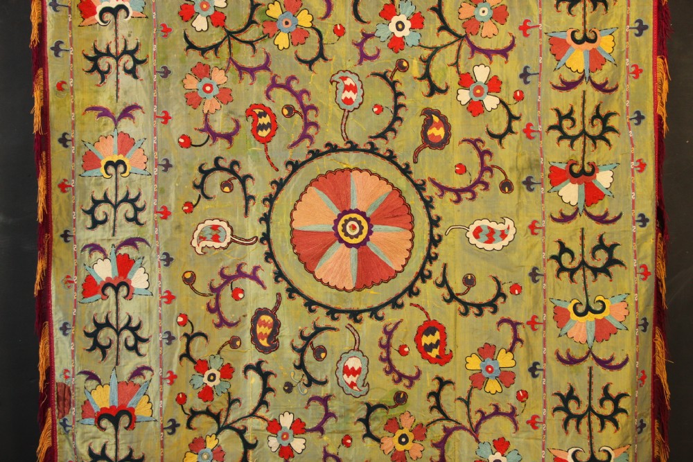 A rectangular green silk embroidered Suzani panel featuring a central circular solar motif flanked by intricate scrolling vines, botanical borders, and fringe edging. View II