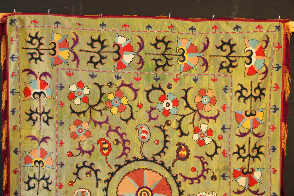 A rectangular green silk embroidered Suzani panel featuring a central circular solar motif flanked by intricate scrolling vines, botanical borders, and fringe edging. View III