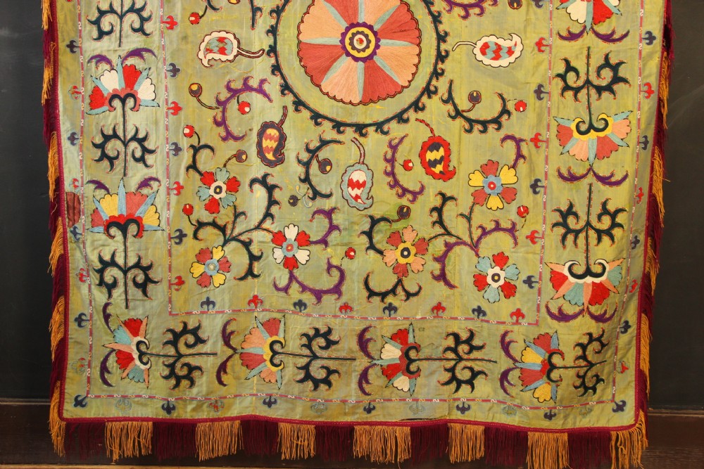 A rectangular green silk embroidered Suzani panel featuring a central circular solar motif flanked by intricate scrolling vines, botanical borders, and fringe edging. View IV