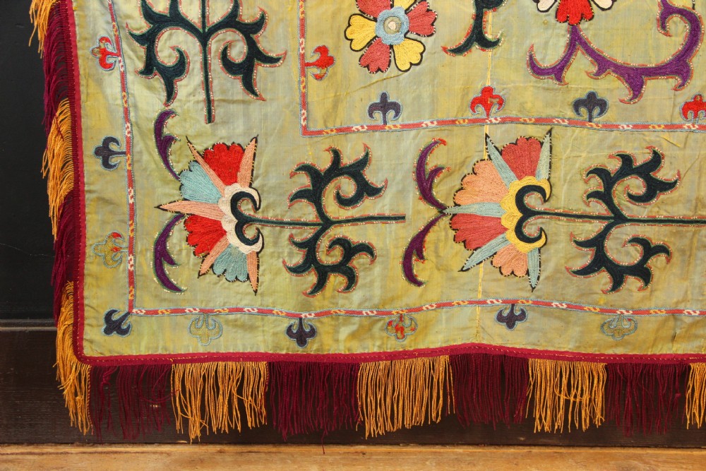 A rectangular green silk embroidered Suzani panel featuring a central circular solar motif flanked by intricate scrolling vines, botanical borders, and fringe edging. View V