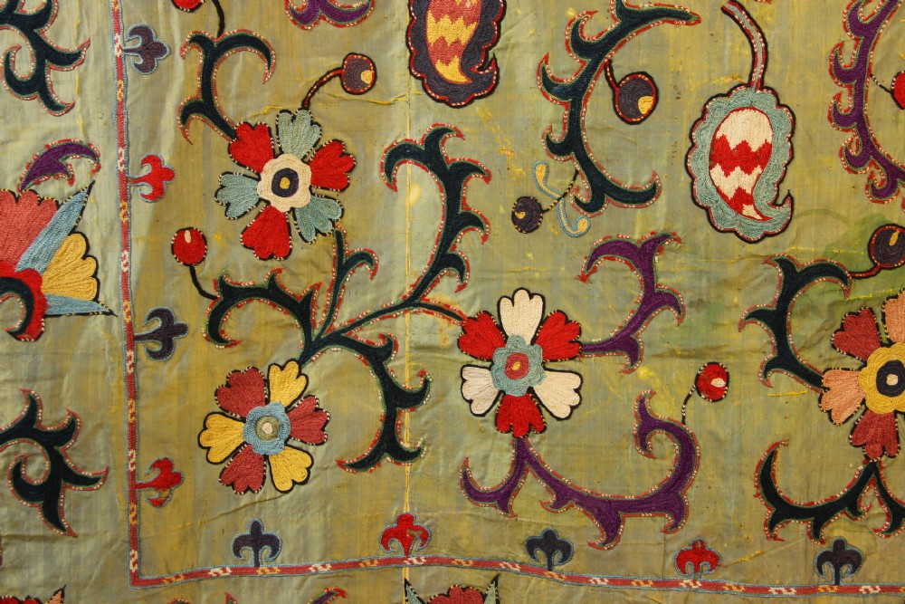 A rectangular green silk embroidered Suzani panel featuring a central circular solar motif flanked by intricate scrolling vines, botanical borders, and fringe edging. View VI