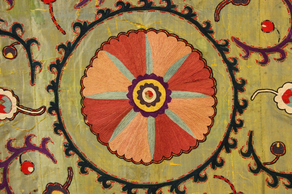 A rectangular green silk embroidered Suzani panel featuring a central circular solar motif flanked by intricate scrolling vines, botanical borders, and fringe edging. View VII