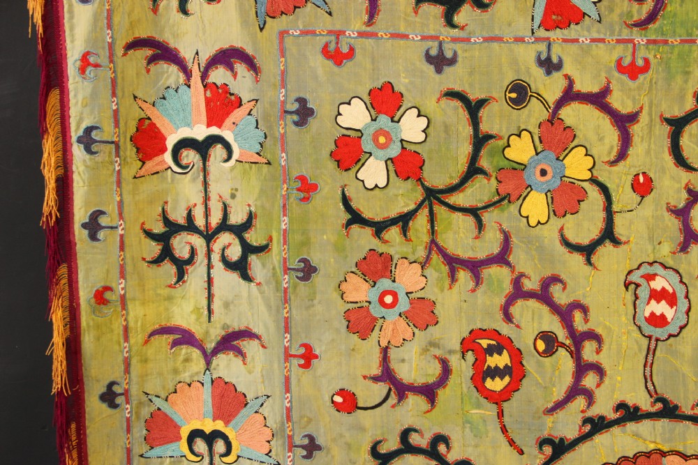 A rectangular green silk embroidered Suzani panel featuring a central circular solar motif flanked by intricate scrolling vines, botanical borders, and fringe edging. View XII