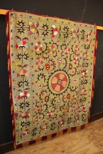 A rectangular green silk embroidered Suzani panel featuring a central circular solar motif flanked by intricate scrolling vines, botanical borders, and fringe edging. View XV