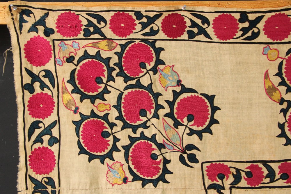 A rectangular hand-embroidered textile featuring central circular pomegranate motifs in crimson silk on a cream ground, framed by a floral border. View V