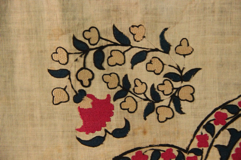 A rectangular hand-embroidered textile featuring central circular pomegranate motifs in crimson silk on a cream ground, framed by a floral border. View X