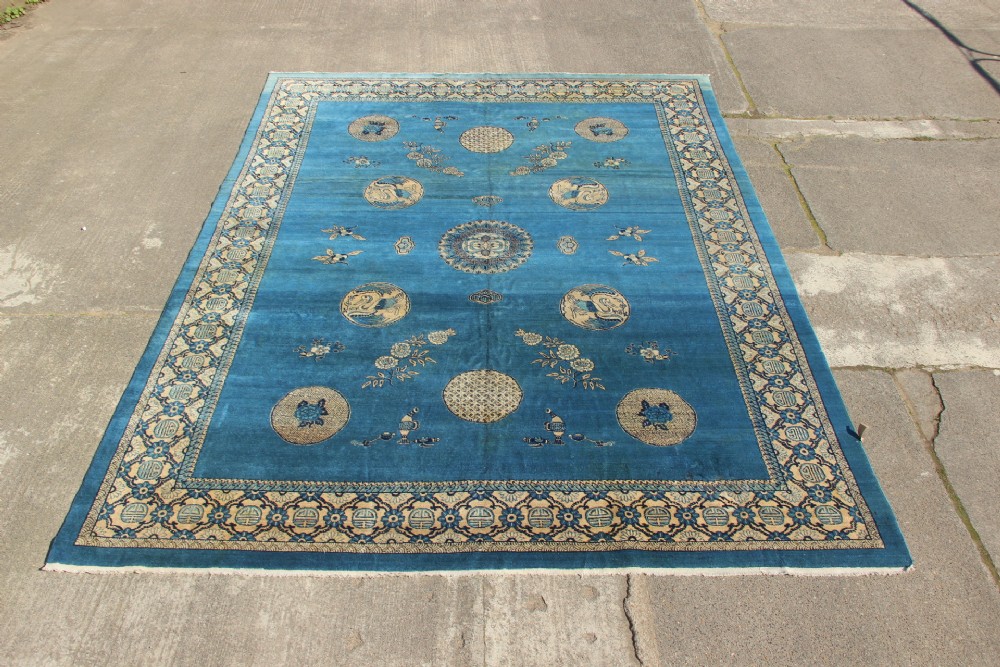 A large rectangular antique Chinese wool carpet featuring a vibrant blue field decorated with circular roundels and floral motifs, enclosed by a wide beige decorative border.