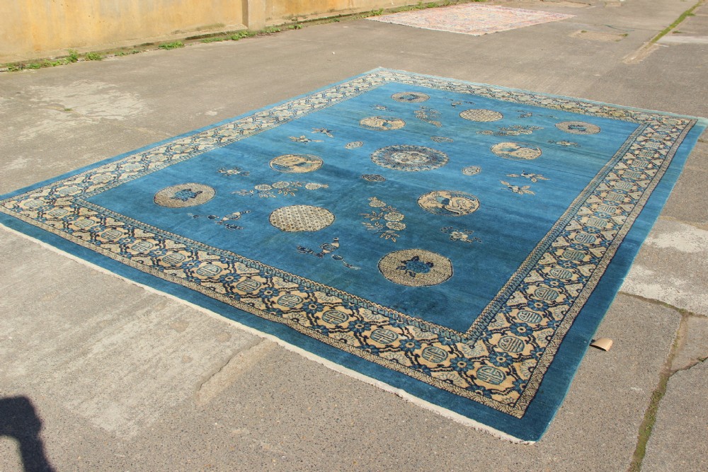 A large rectangular antique Chinese wool carpet featuring a vibrant blue field decorated with circular roundels and floral motifs, enclosed by a wide beige decorative border. View VI