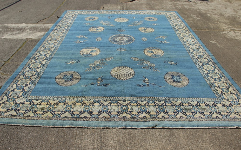 A large rectangular antique Chinese wool carpet featuring a vibrant blue field decorated with circular roundels and floral motifs, enclosed by a wide beige decorative border. View IX