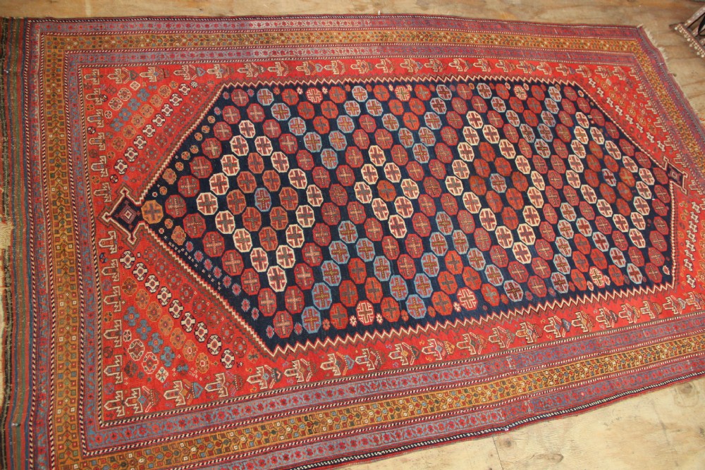 A hand-knotted rectangular wool carpet featuring a dark navy blue hexagonal field adorned with repeating geometric lattice motifs, surrounded by a bold terracotta red border. View IV