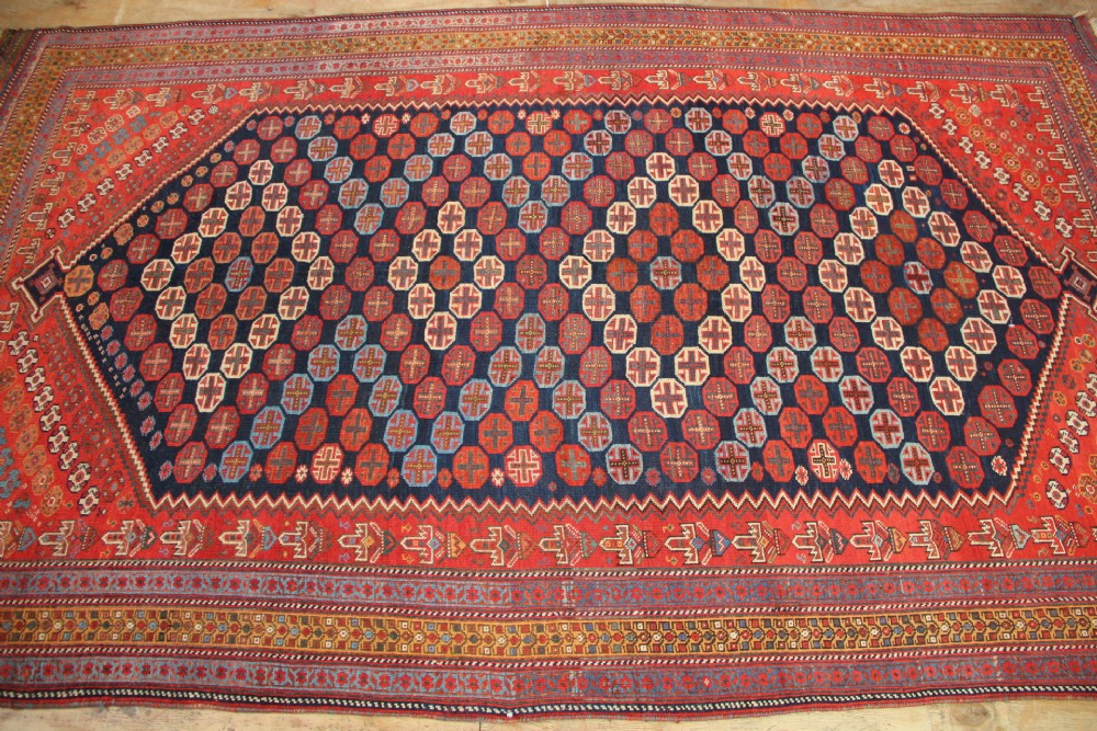 A hand-knotted rectangular wool carpet featuring a dark navy blue hexagonal field adorned with repeating geometric lattice motifs, surrounded by a bold terracotta red border. View V