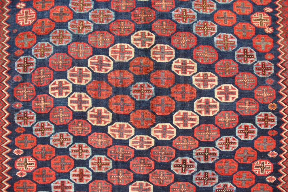 A hand-knotted rectangular wool carpet featuring a dark navy blue hexagonal field adorned with repeating geometric lattice motifs, surrounded by a bold terracotta red border. View VI