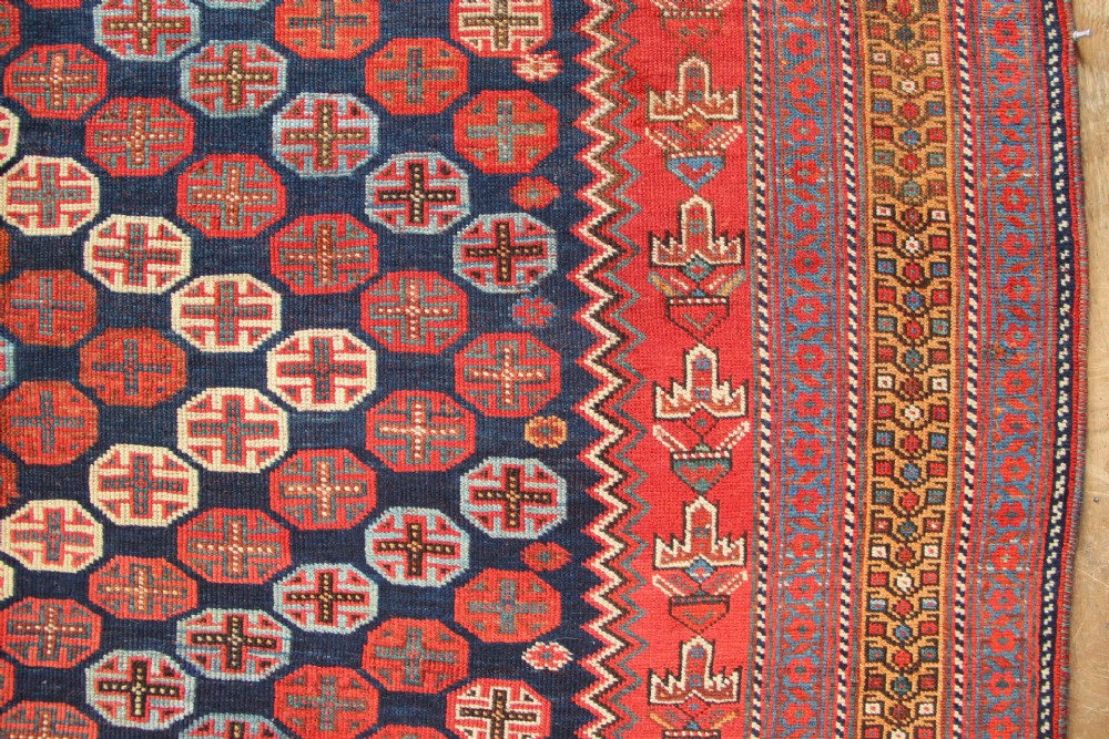 A hand-knotted rectangular wool carpet featuring a dark navy blue hexagonal field adorned with repeating geometric lattice motifs, surrounded by a bold terracotta red border. View VII