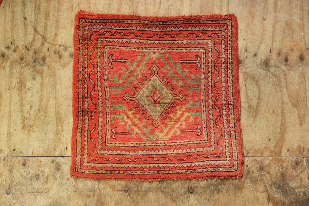 A small square antique hand-knotted wool rug featuring a central diamond medallion surrounded by geometric motifs in terracotta red, sage green, ochre, and dark brown hues.
