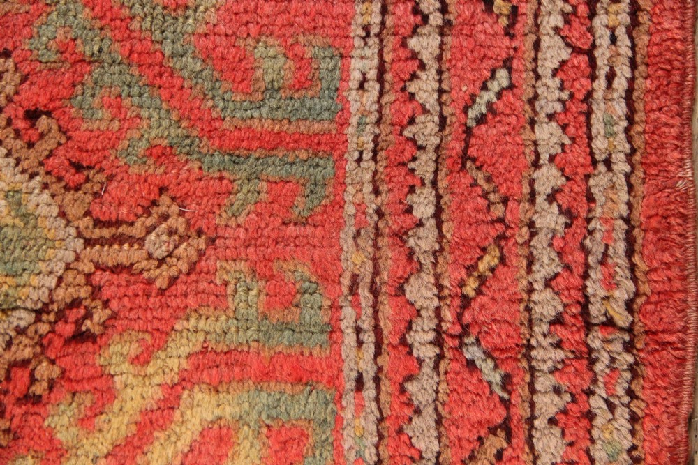A small square antique hand-knotted wool rug featuring a central diamond medallion surrounded by geometric motifs in terracotta red, sage green, ochre, and dark brown hues. View III