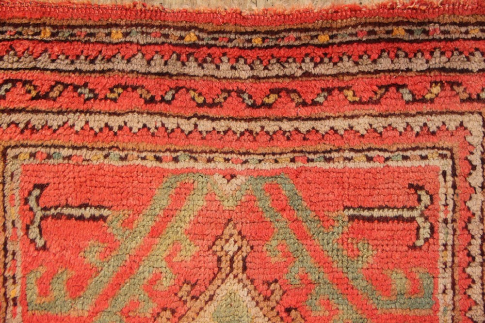 A small square antique hand-knotted wool rug featuring a central diamond medallion surrounded by geometric motifs in terracotta red, sage green, ochre, and dark brown hues. View IV