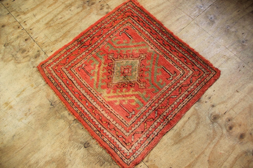A small square antique hand-knotted wool rug featuring a central diamond medallion surrounded by geometric motifs in terracotta red, sage green, ochre, and dark brown hues. View V