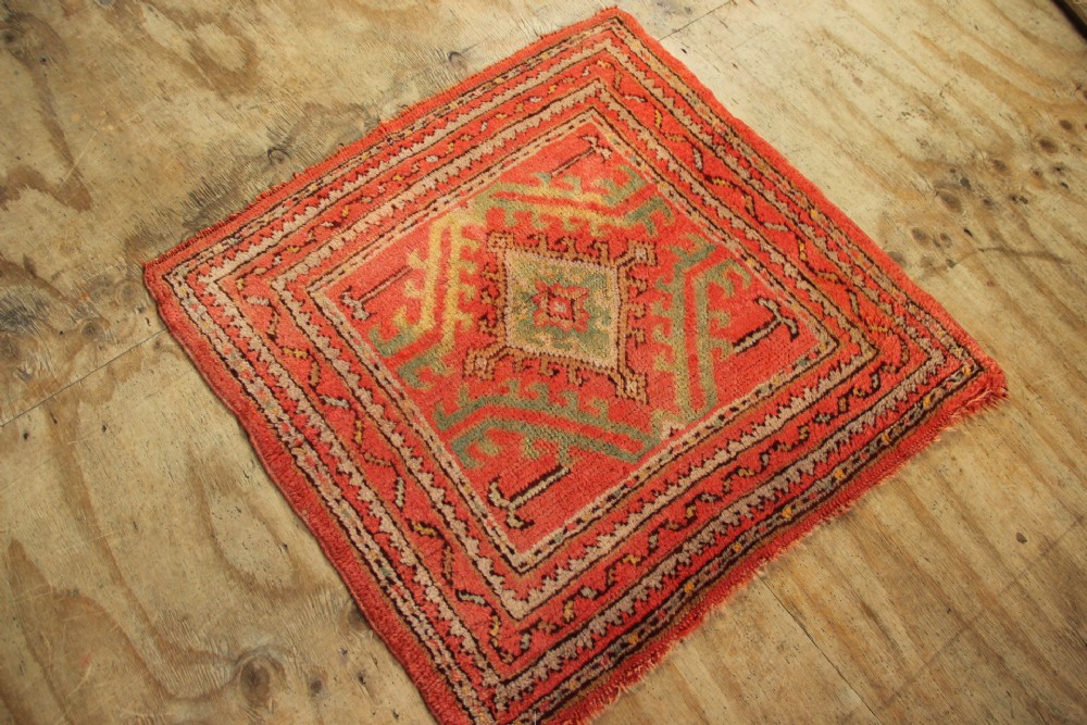 A small square antique hand-knotted wool rug featuring a central diamond medallion surrounded by geometric motifs in terracotta red, sage green, ochre, and dark brown hues. View VI