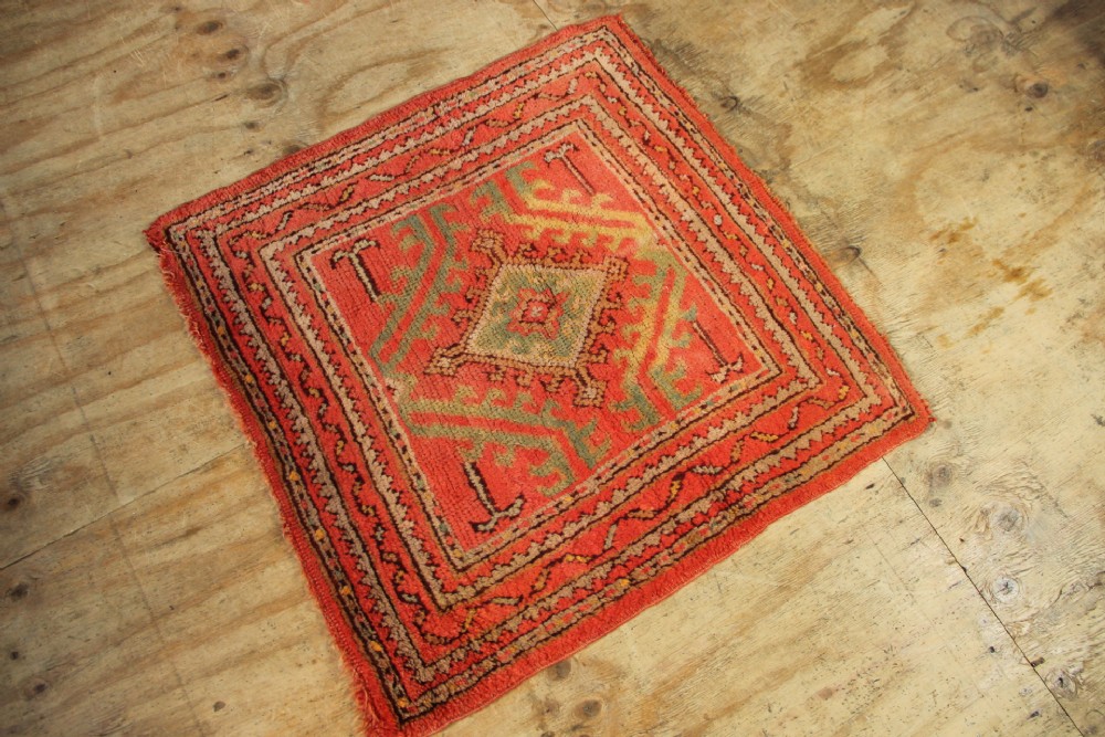 A small square antique hand-knotted wool rug featuring a central diamond medallion surrounded by geometric motifs in terracotta red, sage green, ochre, and dark brown hues. View VII
