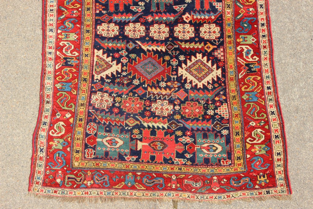 An antique hand-knotted long rug featuring a dark blue central field filled with red, cream, and teal geometric tribal motifs framed by a wide red border. View II