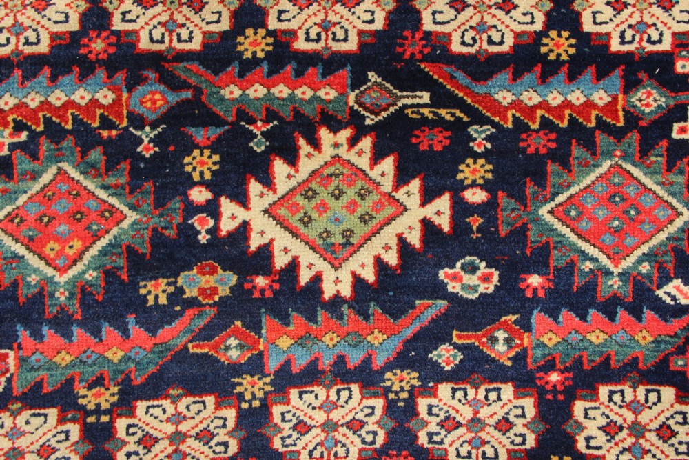 An antique hand-knotted long rug featuring a dark blue central field filled with red, cream, and teal geometric tribal motifs framed by a wide red border. View V