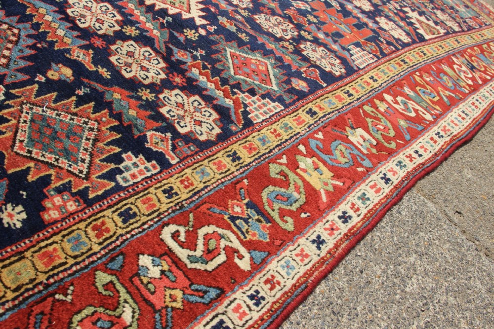 An antique hand-knotted long rug featuring a dark blue central field filled with red, cream, and teal geometric tribal motifs framed by a wide red border. View IX