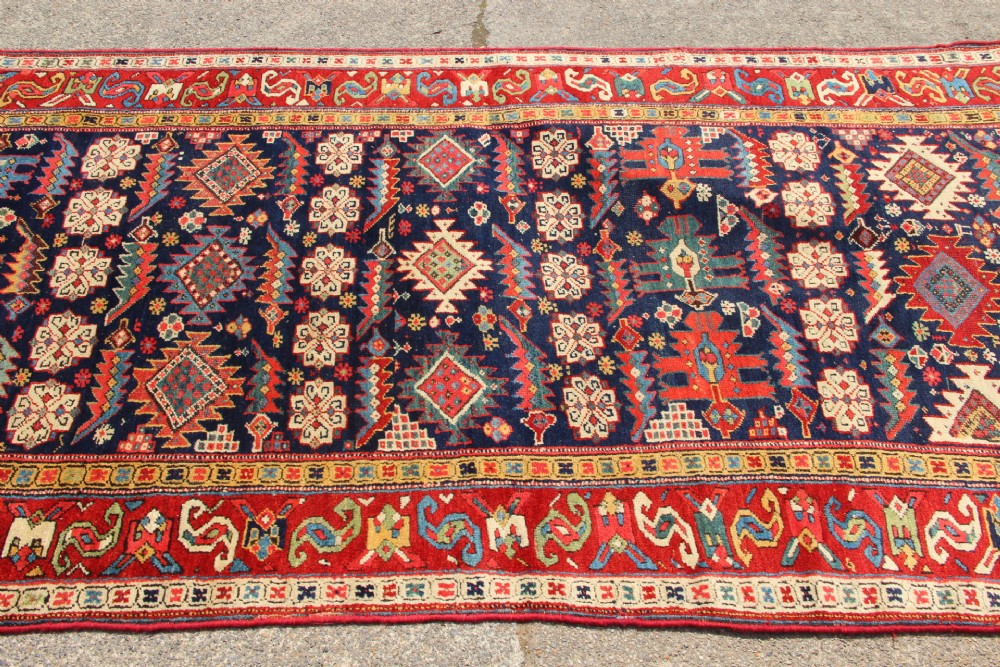 An antique hand-knotted long rug featuring a dark blue central field filled with red, cream, and teal geometric tribal motifs framed by a wide red border. View X