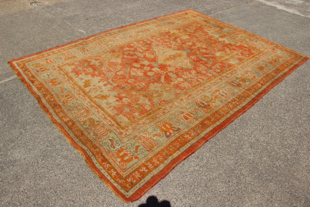 A large hand-woven 19th-century Ghiordes Oushak wool carpet featuring a central diamond medallion, intricate geometric borders, and a terracotta and pale green palette. View X