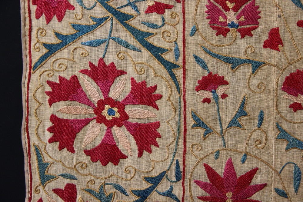 A rectangular hand-embroidered suzani textile featuring crimson floral motifs and blue scrolling vines on a cream ground. View III