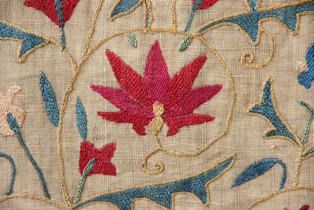 A rectangular hand-embroidered suzani textile featuring crimson floral motifs and blue scrolling vines on a cream ground. View V