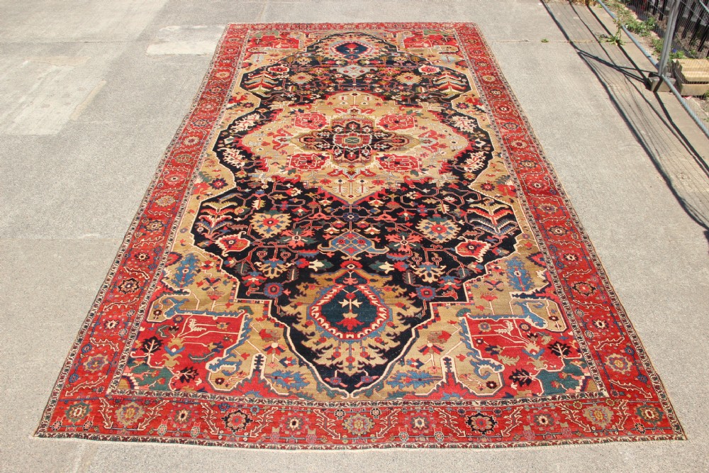 A large hand-knotted Persian Serapi carpet featuring a central geometric medallion in ochre and navy blue, surrounded by a navy field and wide terracotta red border.