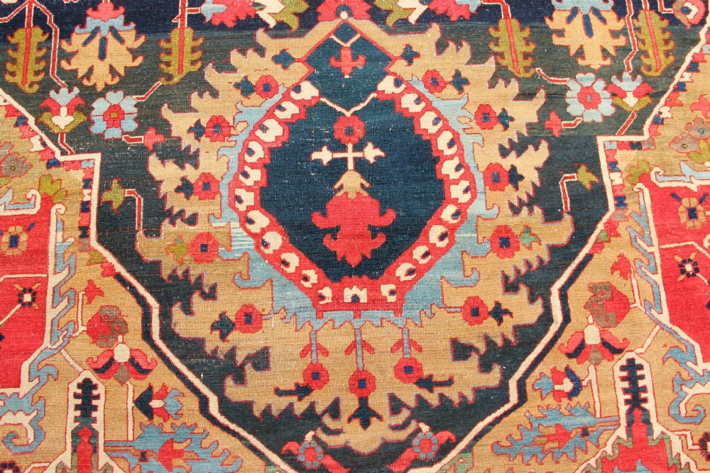 A large hand-knotted Persian Serapi carpet featuring a central geometric medallion in ochre and navy blue, surrounded by a navy field and wide terracotta red border. View III