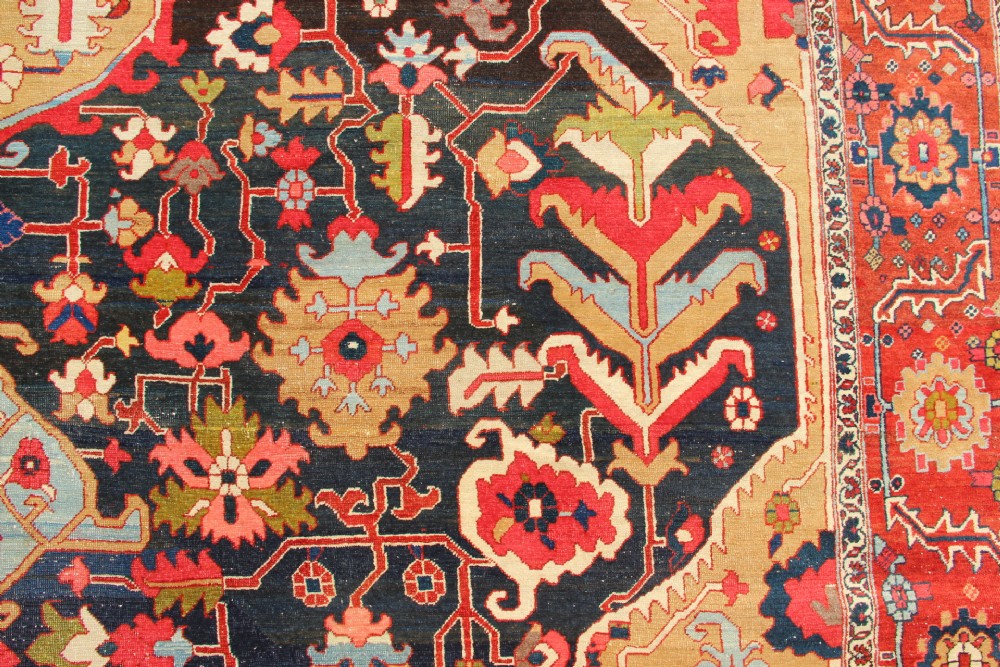 A large hand-knotted Persian Serapi carpet featuring a central geometric medallion in ochre and navy blue, surrounded by a navy field and wide terracotta red border. View IV